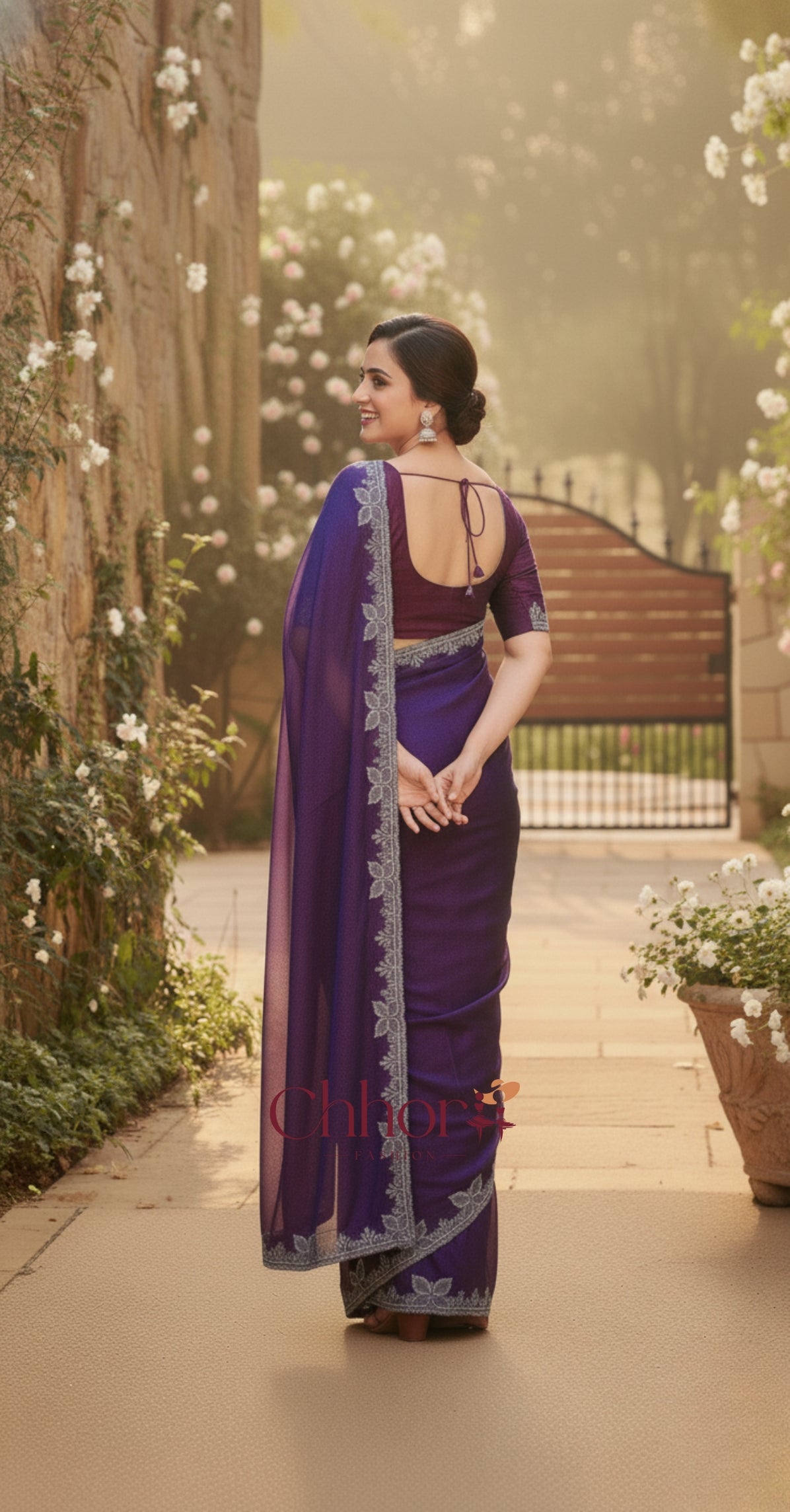 Woman in a purple saree walking along a path with flowers and a gate in the background | Chhorii Fashion